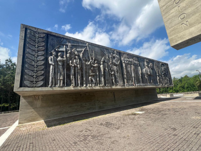 Este muro muestra en tono gris oscuro figuras humanas de aspecto histórico o militar, marchando con banderas, lanzas y a caballo en lo que parece una procesión o gesta heroica. Jorge González