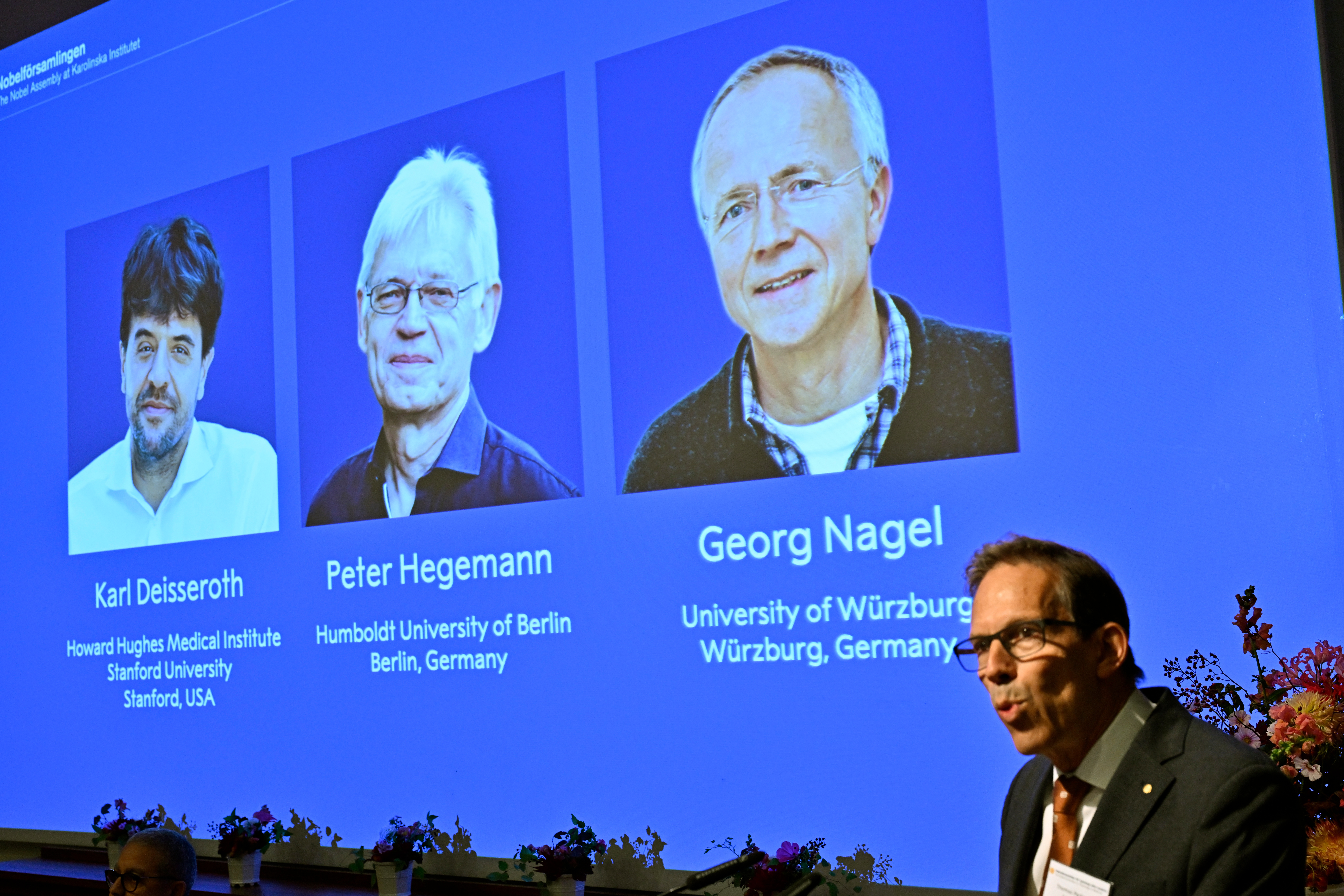 Karl Deisseroth, Peter Hegemann y Georg Nagel se ven en la pantalla en el anuncio de que se les ha concedido el Nobel de Medicina de 2026, en una conferencia de prensa en el Nobel Forum del Instituto Karolinska en Estocolmo, Suecia, el 5 de octubre de 2026. (Pontus Lundahl/TT News Agency via AP)
